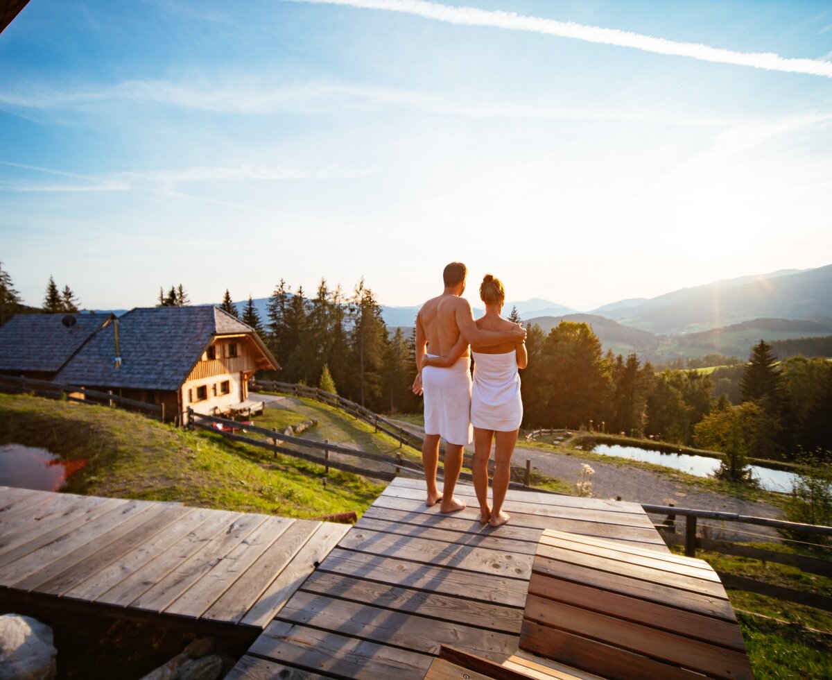 Wellness auf der Alm, Almchalet Nikola in der Steiermark | © Urlaub am Bauernhof / Daniel Gollner Ein paar mit Handtüchern um den Körper stehen nach der Sauna am Holzsteg und blicken in die Ferne, im Hintergrund befindet sich die Almhütte | © Urlaub am Bauernhof / Daniel Gollner