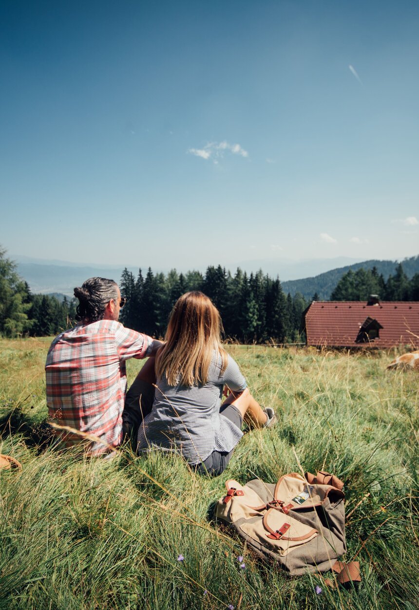Ein Paar sitzt mit dem Rücken zum Betrachter im Graz, hinter ihnen liegen die Rucksäcke, sie blicken auf die Almhütte, von der das Dach zu sehen ist, eine Kuh liegt etwas entfernt vor ihnen im Gras, mit Blick auf die Aussicht | © Urlaub am Bauernhof / Daniel Gollner