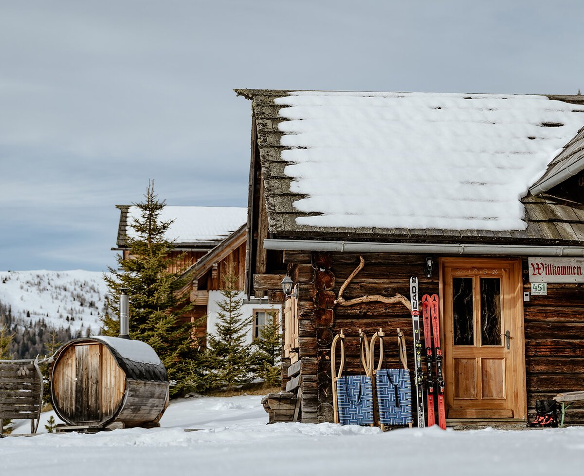 Eine urige Almhütte aus Holz in schneebedeckter Landschaft. Vor der Hütte lehnen Ski und Schlitten. Links davon ist eine Sauna aus Holz zu sehen und im Hintergrund sieht man eine weitere Holzhütte. | © Urlaub am Bauernhof Steiermark / (c) Wolfgang Spekner 