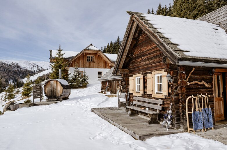 Idyllisch gelegene Almhütte in einer Winterlandschaft. Vor der Holzhütte lehnen Schlitten und links im Bild sieht man eine Fassauna. | © Urlaub am Bauernhof Steiermark | (c) Wolfgang Spekner