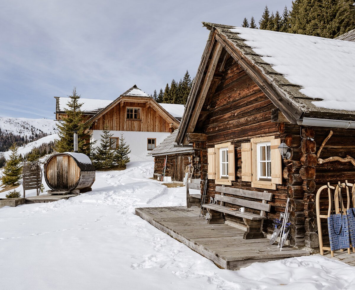 Idyllisch gelegene Almhütte in einer Winterlandschaft. Vor der Holzhütte lehnen Schlitten und links im Bild sieht man eine Fassauna. | © Urlaub am Bauernhof Steiermark | (c) Wolfgang Spekner