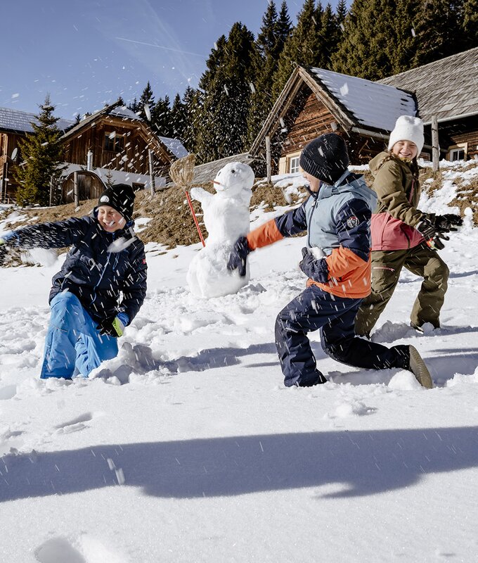 Zwei Jungen und ein Mädchen bewerfen sich auf einer Wiese mit Schnee mit Schneebällen. Dahinter stehen zwei Almhütten aus Holz. | © Urlaub am Bauernhof Steiermark | (c) Wolfgang Spekner