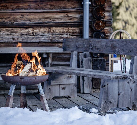 In der Mitte einer Holzsitzgarnitur steht eine Feuerschale, in der Holz brennt. Dahinter ist eine weiße Winterlandschaft.  | © Urlaub am Bauernhof Steiermark | (c) Wolfgang Spekner