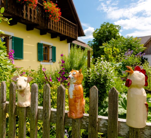 sommerlicher Bauerngarten mit Salat, Kräutern und Gemüse, umgeben von einem Holzzaun, auf dem 3 Keramikfiguren stecken, 1 Kuh, 1 Katze und 1 Hahn, dahinter ist das gelbe Bauernhaus mit Holzbalkon zu sehen | © Urlaub am Bauernhof Steiermark / Wolfgang Spekner