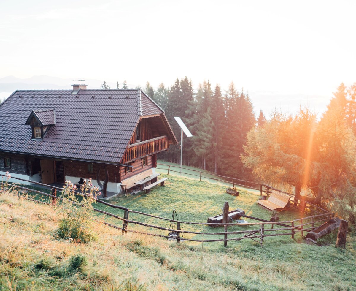 Gesamtansicht einer Almhütte aus Holz mit der Wiese rundherum, mit einem Holzzaun eingezäunt und in das warme Licht des Sonnenaufgangs getaucht. | © Urlaub am Bauernhof Steiermark / Daniel Gollner Almhütte Abfalterer, Steiermark | © Urlaub am Bauernhof Steiermark / Daniel Gollner