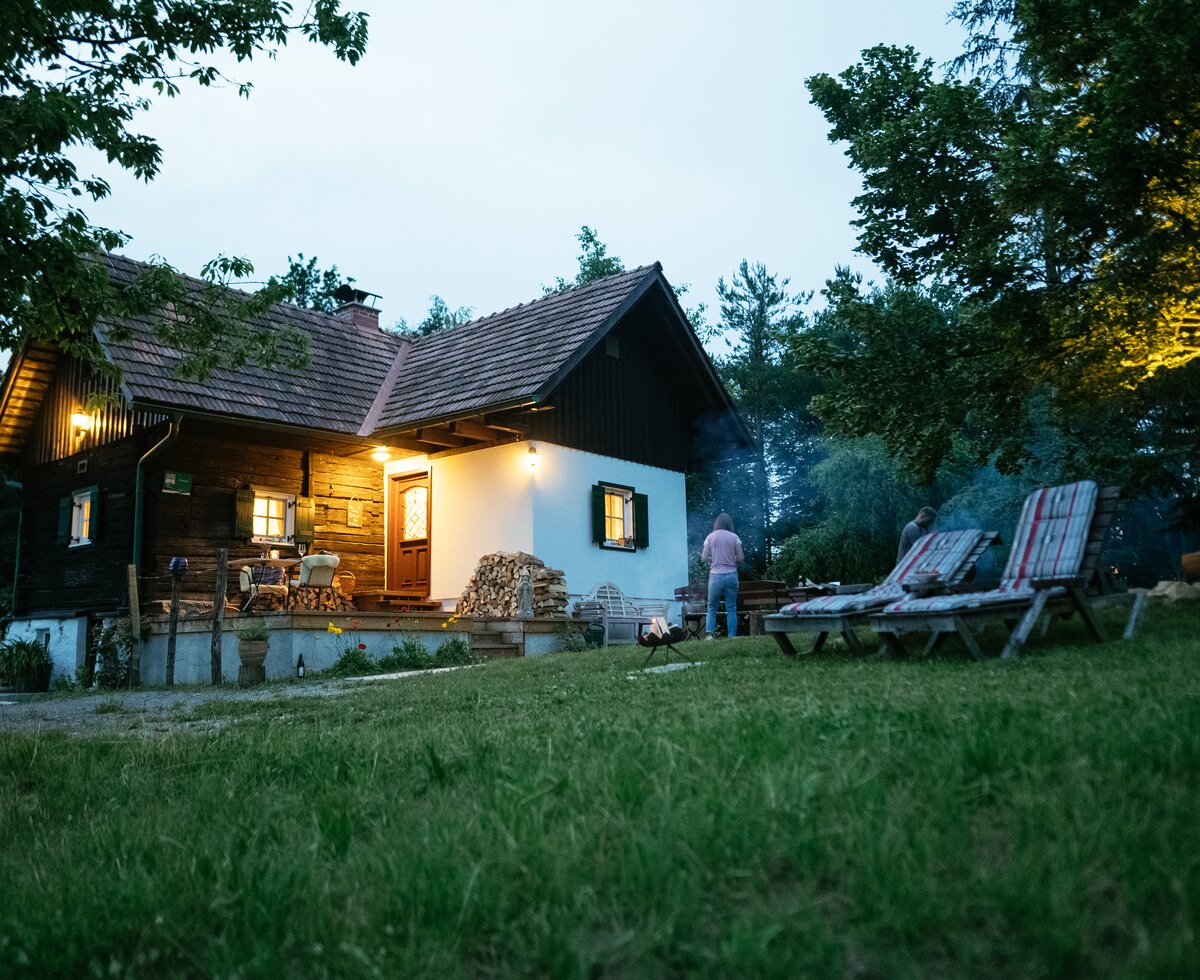 Waldhütte Adam in der Südsteiermark, Steiermark | © Urlaub am Bauernhof Steiermark / Daniel Gollner Die Waldhütte hat ein gemauertes Fundament, darüber besteht sie aus Holz, sie ist in der Dämmerung zu sehen mit erleuchteten Fenstern und im Vordergrund stehen zwei Liegestühle | © Urlaub am Bauernhof Steiermark / Daniel Gollner