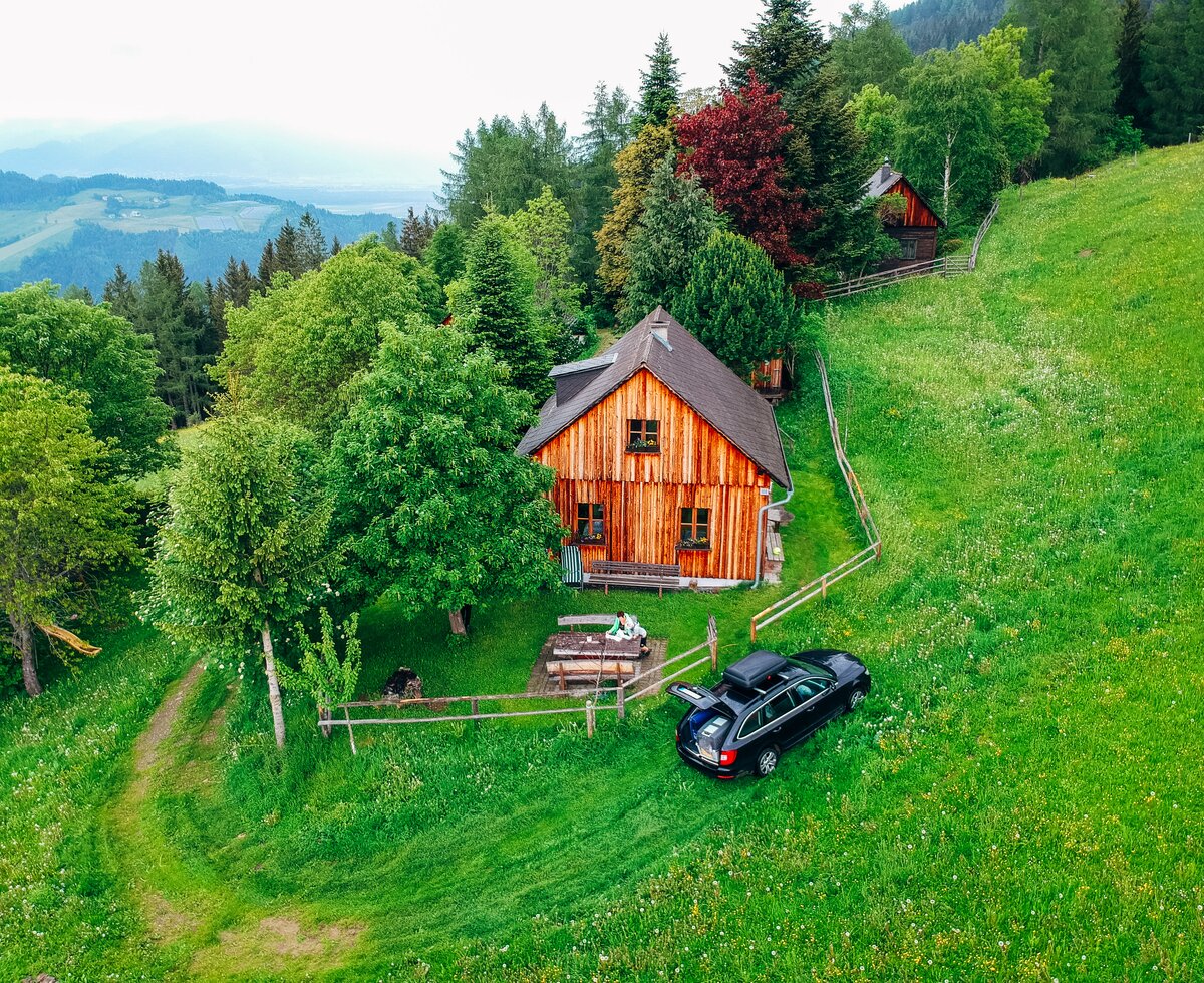 Almhütte Laikam, Steiermark | © Urlaub am Bauernhof Steiermark / Daniel Gollner Luftaufnahme der Almhütte Laikam, die allein neben einem Wald liegt, vor der Hütte steht ein Auto, auf der Holzgarnitur sitzt eine Frau mit grüner Jacke | © Urlaub am Bauernhof Steiermark / Daniel Gollner