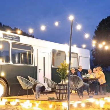 Zu einem Glampingbus umgebauter Grazer Linienbus mit Terrasse, auf der mehrere Personen am Abend mit Beleuchtung sitzen, ein schwarz-weißer Hund geht über die Terrasse. | © Bauernhof Lutzmann / Katrin Lutzmann