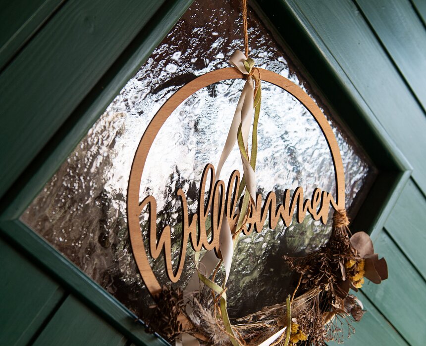 Auf einer grünen Tür hängt ein Türkranz aus Trockenblumen mit einem aus Holz geschnitzten Willkommen in der Mitte. | © Urlaub am Bauernhof / Carletto Ferrari-Brunnenfeld 