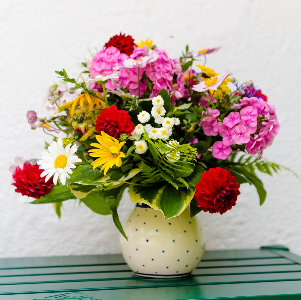 Eine weiße, bauchige Blumenvase mit grünen Punkten steht auf einem grün lackierten Tisch vor weißen Hintergrund, darin ein üppiger Strauß mit roten, gelben, weißen, rosa Sommerblumen aus dem Garten | © Urlaub am Bauernhof Steiermark / Wolfgang Spekner