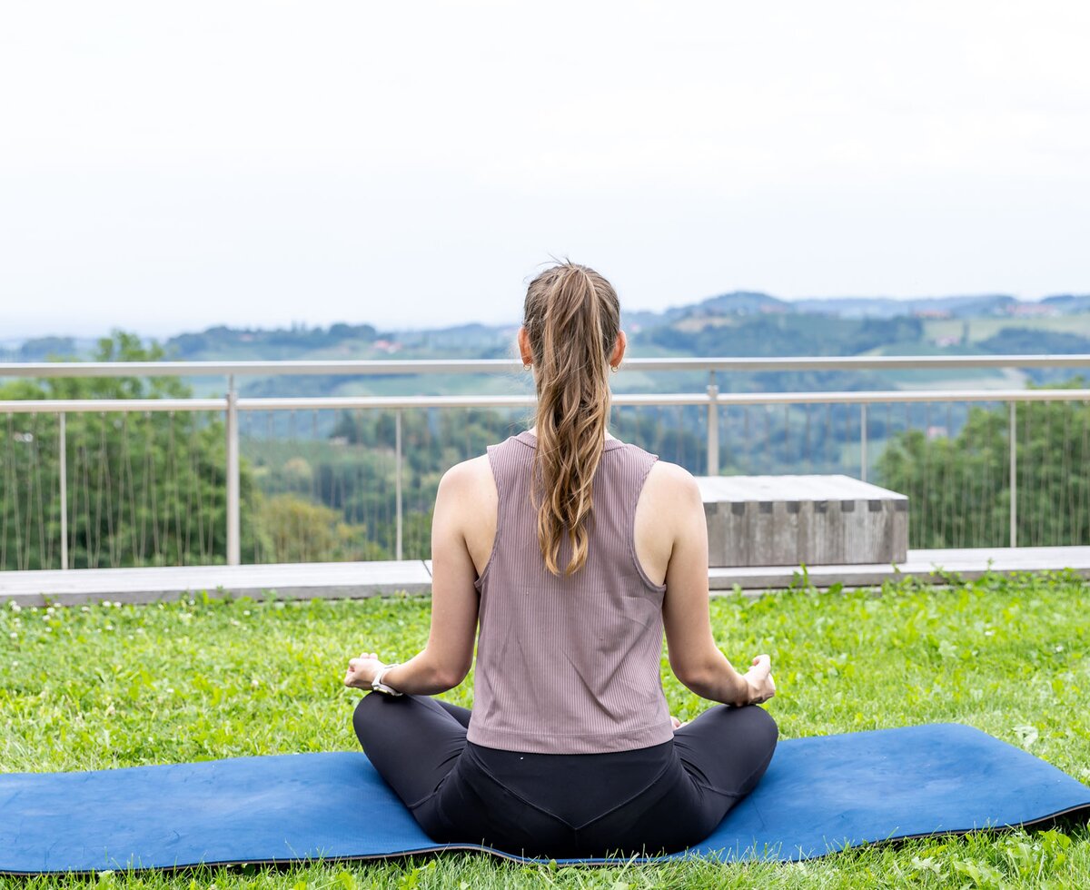 Eine Frau sitzt im Yogasitz im Garten auf der Matte und blickt auf die Weinhänge | © Urlaub am Winzerhof / Elisabeth Fröhlich Eine Frau beim Yoga im Weingarten | © Urlaub am Winzerhof / Elisabeth Fröhlich