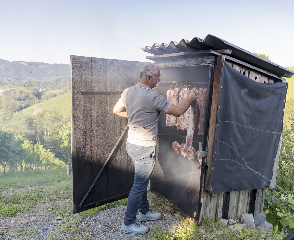 Beim Fleischräuchern am Winzerhof in der Südsteiermark | © Urlaub am Bauernhof / Elisabeth Fröhlich Am oberen Ende des Weinhangs steht eine Holzhütte, in der das Fleisch zum Räuchern aufgehängt wird. Ein Mann steht vor der Hütte und hängt das Fleisch auf, es raucht aus der Hütte. | © Urlaub am Bauernhof / Elisabeth Fröhlich