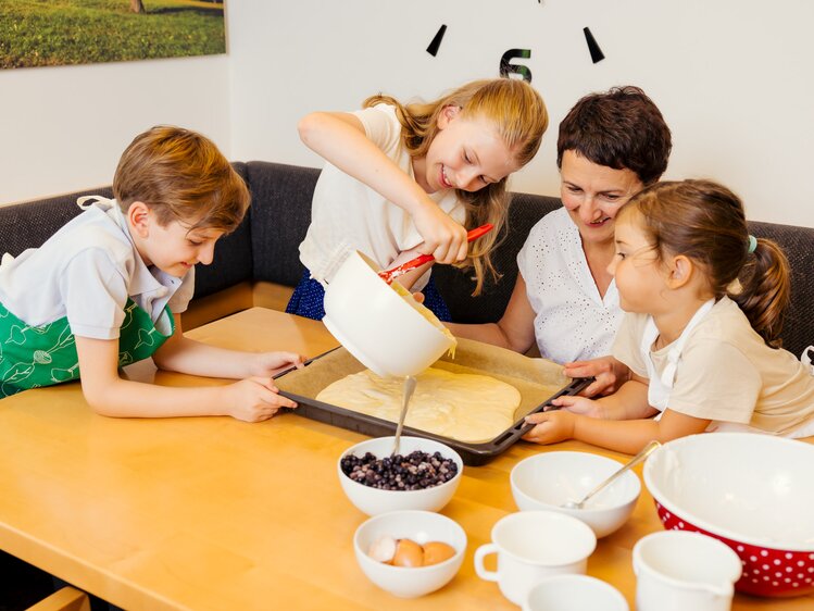 Ein Mädchen gießt unter den wachsamen Augen der Bäuerin den Kuchenteig auf ein Backblech, ein Bub und ein Mädchen schauen zu und halten das Blech | © UaB Steiermark | Wolfgang Spekner