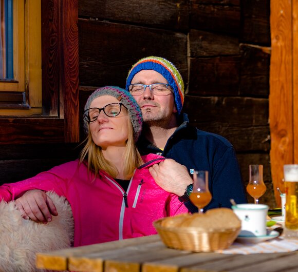 Ein Paar sitzt im Winter an der Hauswand und genießt die Sonne, Urlaubsregion Murau-Murtal, Steiermark | © Urlaub am Bauernhof Steiermark / Wolfgang Spekner