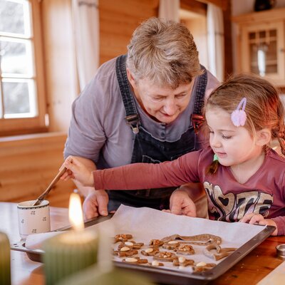 Mädchen backt gemeinsam mit Oma Weihnachtskekse in einer Bauernstube. Im Vordergrund sieht man einen Adventkranz, auf dem zwei Kerzen brennen. | © UaB Steiermark / Wolfgang Spekner