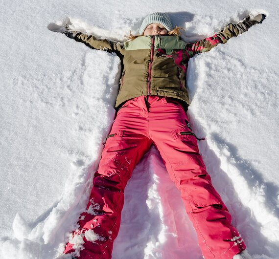 Ein Kind mit rosa Skihose und braunem Anorak liegt im Schnee und macht einen Schneeengel | © Urlaub am Bauernhof Steiermark / Wolfgang Spekner