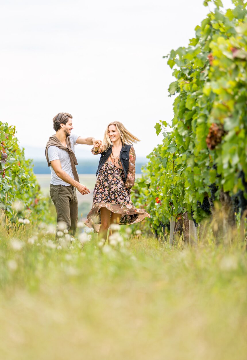 Ein junges Paar ist in einem Weinberg beim Tanzen und hat sichtlich Spaß. Ihre Kleidung und Frisuren deuten darauf hin, dass es sich um eine Freizeit-Aktivität handelt.