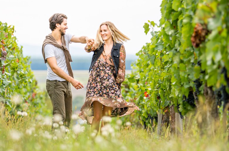 Ein junges Paar ist in einem Weinberg beim Tanzen und hat sichtlich Spaß. Ihre Kleidung und Frisuren deuten darauf hin, dass es sich um eine Freizeit-Aktivität handelt.