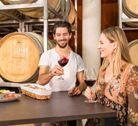 Ein Paar trinkt Wein in einer Weinkellerei umgeben von Holzfässern. Vor ihnen liegen verschiedene Leckereien auf einem dunklen Holztisch.