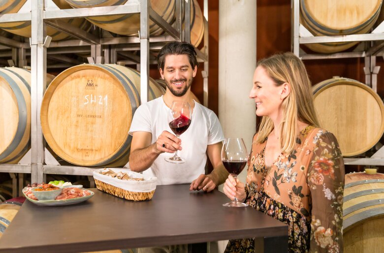 Ein Paar trinkt Wein in einer Weinkellerei umgeben von Holzfässern. Vor ihnen liegen verschiedene Leckereien auf einem dunklen Holztisch.