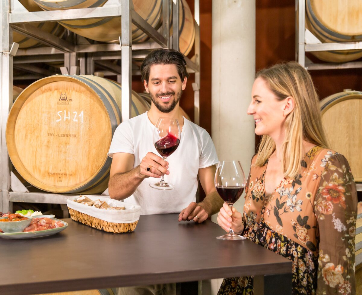 Ein Paar trinkt Wein in einer Weinkellerei umgeben von Holzfässern. Vor ihnen liegen verschiedene Leckereien auf einem dunklen Holztisch.