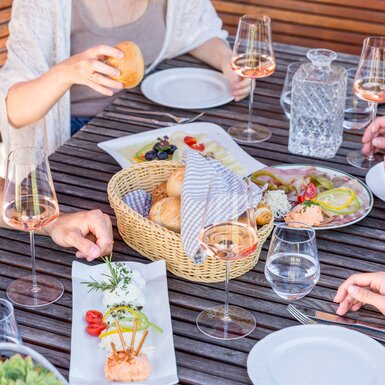 Eine Gruppe genießt ein luxuriöses Picknick mit Köstlichkeiten auf einer Terrasse. Auf dem Holztisch stehen Weinkelche, eine Brotkorb und eine kreative Präsentation von Appetithäppchen.