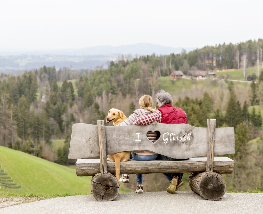 Ein älteres Paar sitzt auf einer Holzbank, das sie mit einem Hund teilen. Sie blicken auf eine bergige Landschaft mit Wald und Wiesen.