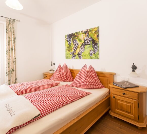 Gemütliches Hotelzimmer mit Holzmöbeln, Karo-Bettwäsche in Rot und Weiß sowie einem Bild von Trauben an der Wand. Der Blick geht durch ein Fenster auf eine Terrasse.