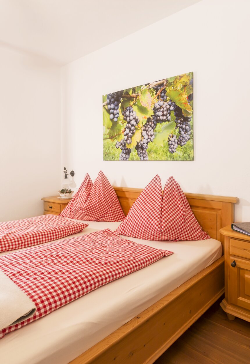 Gemütliches Hotelzimmer mit Holzmöbeln, Karo-Bettwäsche in Rot und Weiß sowie einem Bild von Trauben an der Wand. Der Blick geht durch ein Fenster auf eine Terrasse.