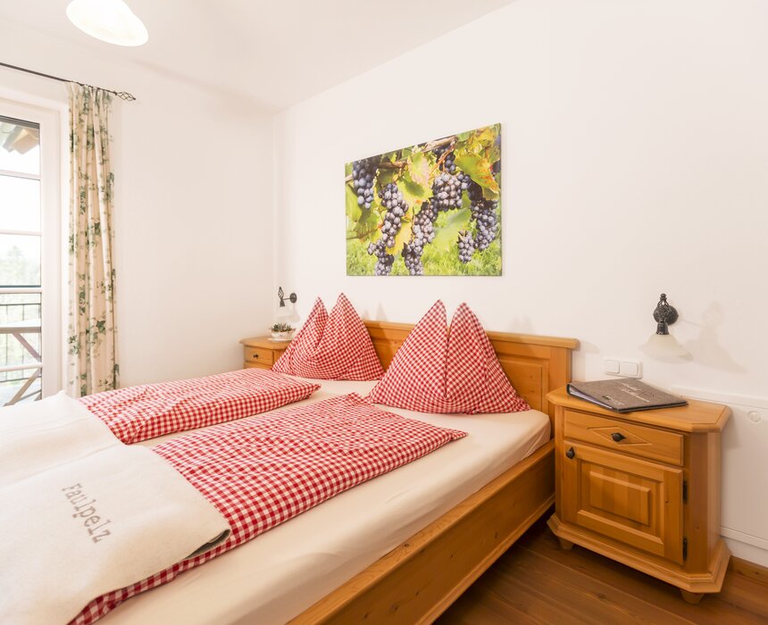 Gemütliches Hotelzimmer mit Holzmöbeln, Karo-Bettwäsche in Rot und Weiß sowie einem Bild von Trauben an der Wand. Der Blick geht durch ein Fenster auf eine Terrasse.
