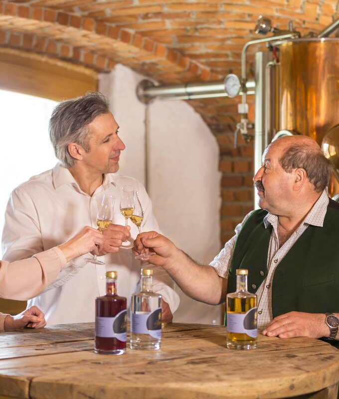 Drei Personen stoßen mit Gläsern an und haben eine gesellige Unterhaltung in einer rustikalen Brennerei mit Kupferkesseln und Ziegelwänden.