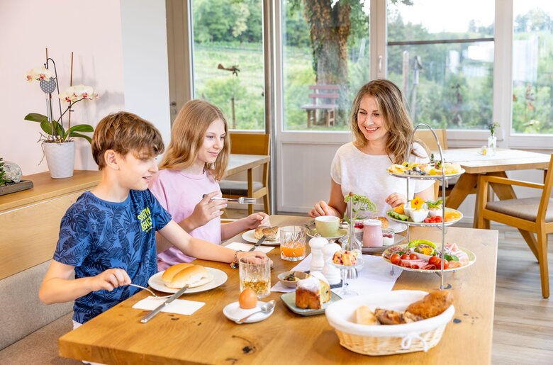 Eine Familie, die an einem reich gedeckten Tisch mit Brot, Obst, Gebäck und anderen Speisen sitzt und in einem gemütlichen Esszimmer mit Blick auf eine Gartenlandschaft isst.