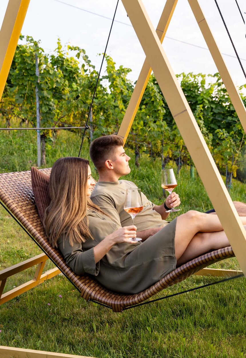 Ein junges Paar ruht sich in einem Hängestuhl aus und genießt Getränke, umgeben von einem sommerlichen Weinberg.