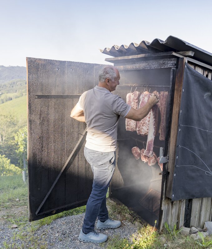 Ein älterer Mann hängt rohe Fleischstücke in einer hölzernen Raucherkabine auf. Im Hintergrund ist eine grüne Landschaft zu sehen.
