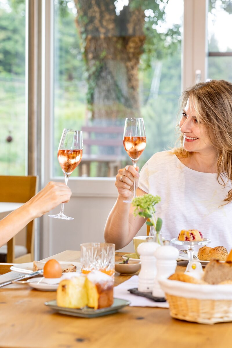 Zwei Frauen sitzen an einem gedeckten Tisch und genießen einen Snack mit Getränken in einem hellen, gemütlichen Ambiente mit Blick ins Grüne.