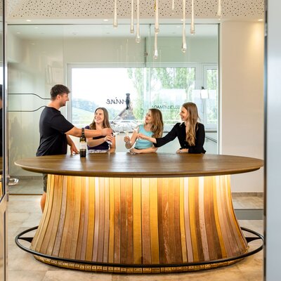 Im Bild sind vier Personen zu sehen, die sich an einem runden Holztisch in einer modern eingerichteten Umgebung unterhalten. Über dem Tisch hängt eine dekorative Beleuchtung. Die Atmosphäre wirkt entspannt und einladend. | © Urlaub am Bauernhof / Elisabeth Fröhlich