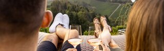 Zwei Personen sitzen auf einer Terrasse mit Blick auf eine hügelige Landschaft. Sie halten Weingläser in den Händen und genießen offenbar einen entspannenden Moment. Die Beine der Personen sind sichtbar, sie tragen legere Sommerbekleidung.