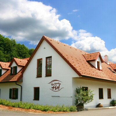Die Ansicht des Bauernhofs mit weißer Fassade und terrakottafarbenen Ziegeldächern, eingebettet in die grüne Landschaft.