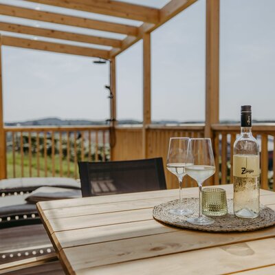 Private Terrasse der Ferienwohnung "Fernblick" mit Holztisch, Wein, Sonnenliegen und Blick auf die Hügellandschaft.