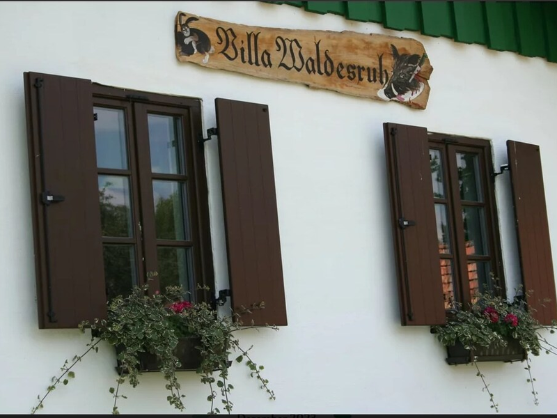 Die weiße Fassade des Hofes mit zwei Fenstern, offenen braunen Fensterläden, Blumenkästen und dem Holzschild "Villa Waldesruh".
