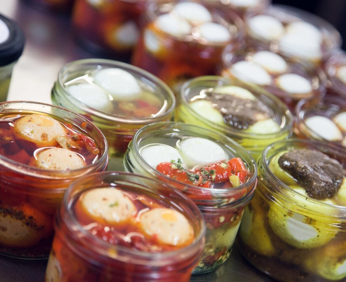 Eingemachte Spezialitäten in Gläsern, darunter Frischkäse-Kugeln und eingelegtes Gemüse, erhältlich im Ferienhaus.