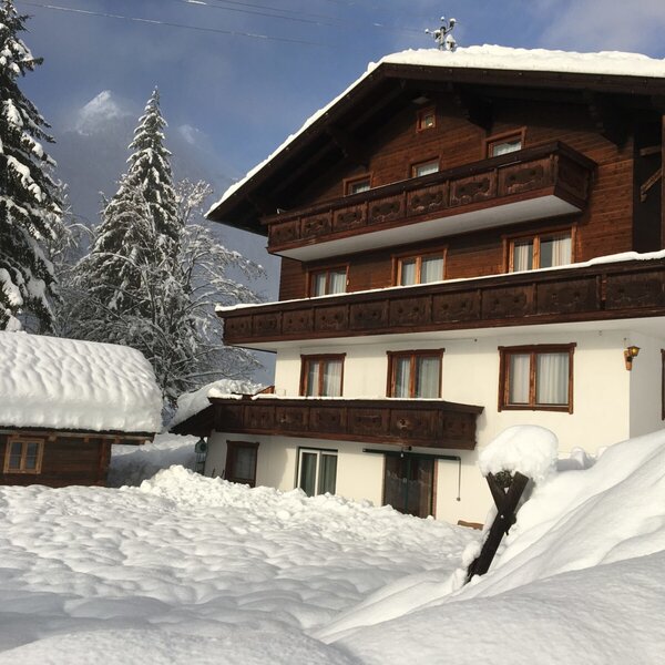 Die Alm-Appartements Regina, bestehend aus einem mehrstöckigen Gebäude mit Balkonen und einem kleineren Holzhaus, sind komplett von winterlichem Schnee bedeckt.