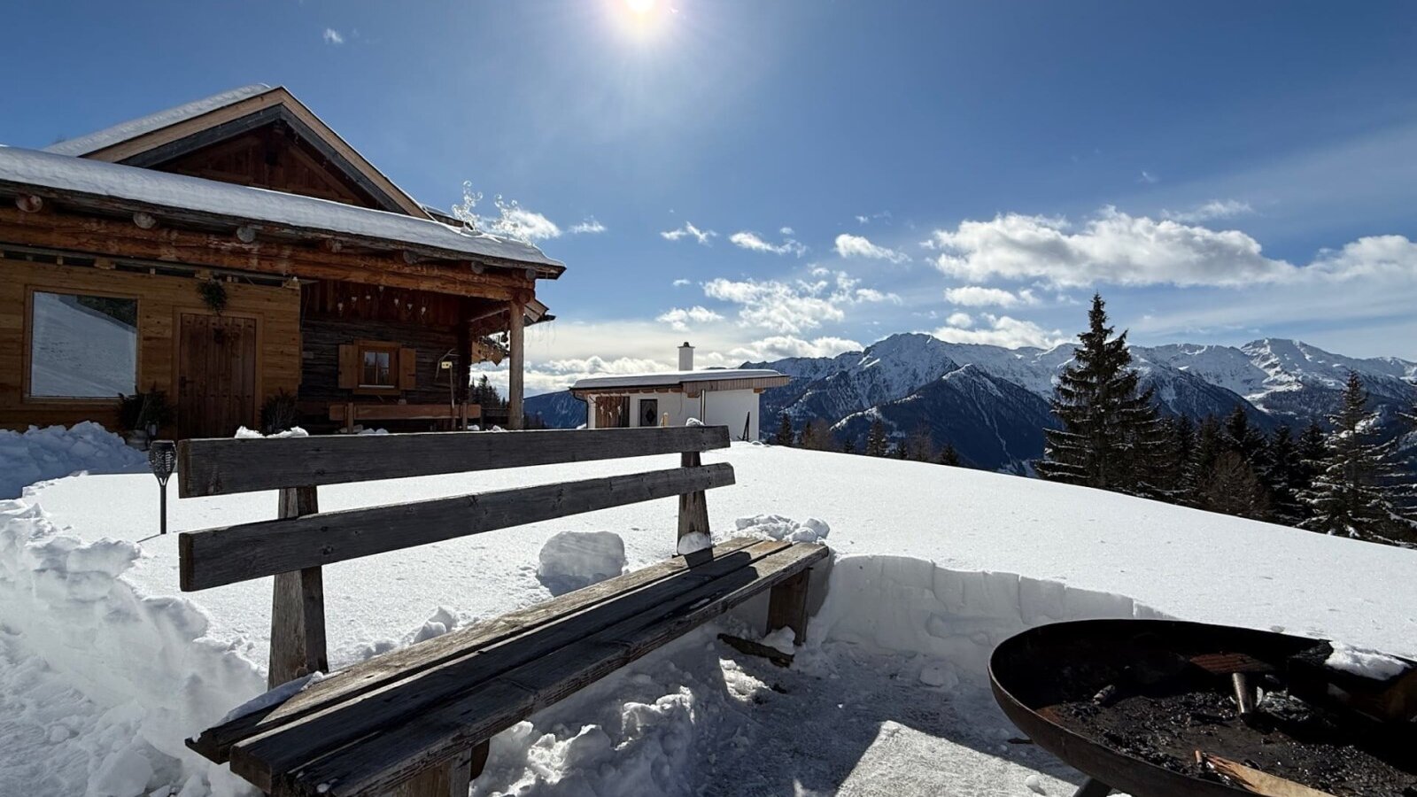 Die Alm mit einer Holzbank und Feuerstelle im verschneiten Außenbereich, mit Bergblick.