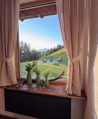Blick aus dem Fenster der Alm auf die grüne Hügellandschaft mit Bäumen und Bergen.