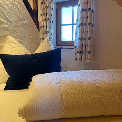 Ein gemütlicher Schlafbereich auf der Alm mit weichem Bettzeug und einem urigen kleinen Fenster mit gemusterten Vorhängen.