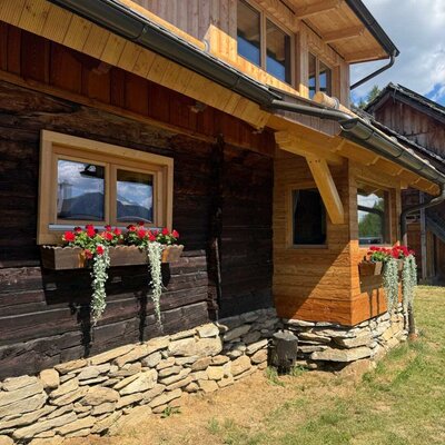 Die charmante Außenansicht der Alm mit traditionellen Holzelementen, Natursteinsockel und blühenden Blumenkästen.