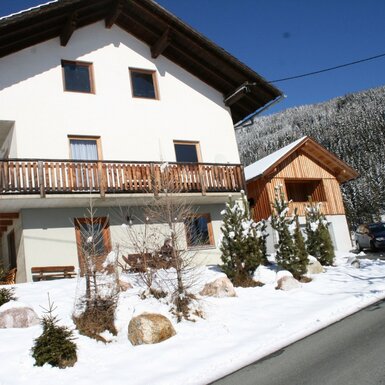 Die Alm mit weißer Fassade, Holzbalkon und angrenzendem Holzgebäude, umgeben von Schnee und einer verschneiten Berglandschaft.