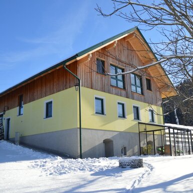 Der Hof im Winter mit schneebedeckter Umgebung, gelber Fassade, Holzverkleidung und einem überdachten Anbau.