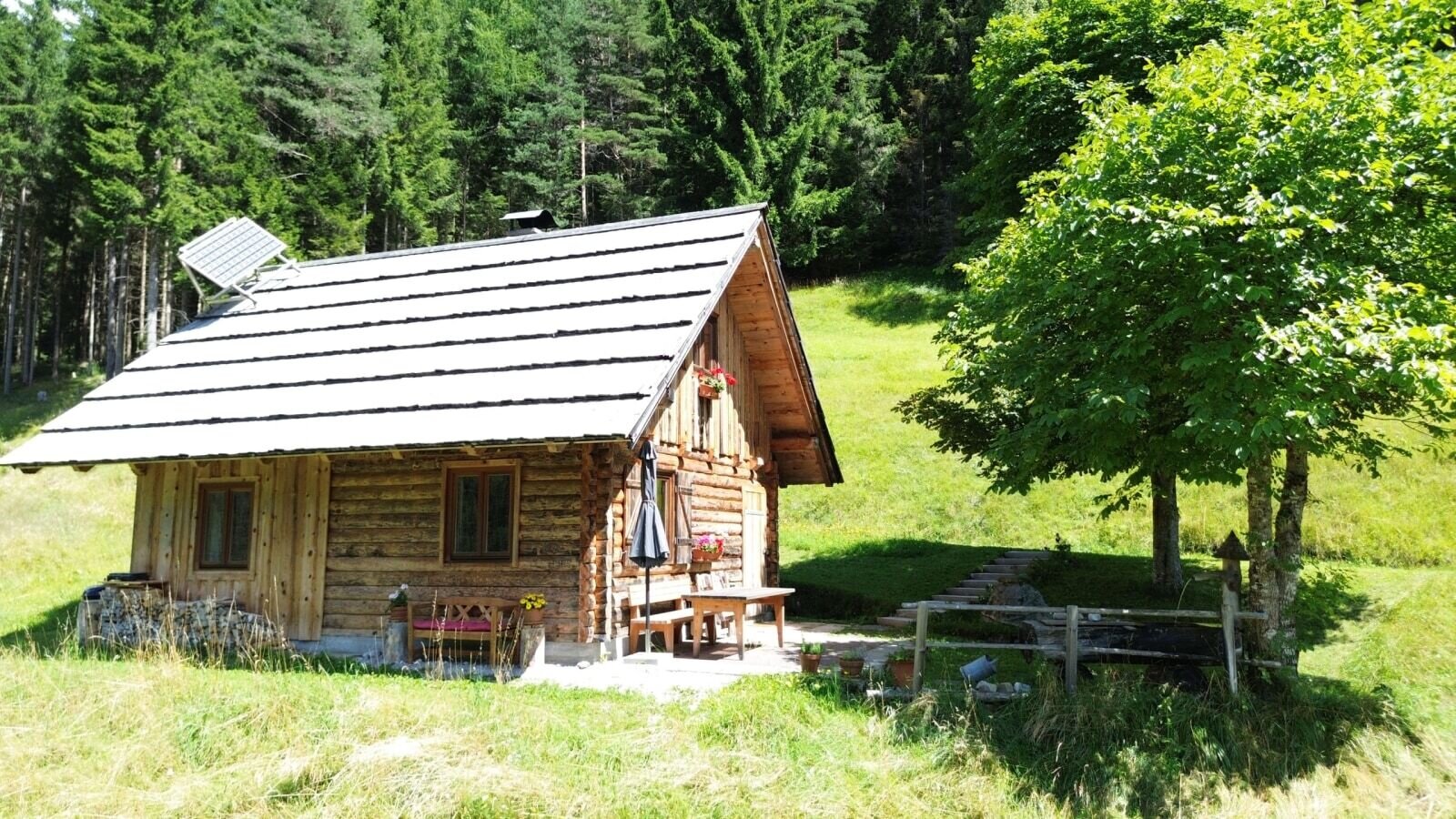 Almhütte mit Solarpaneel und einem Sitzbereich im Freien, umgeben von Wiesen und Wald.