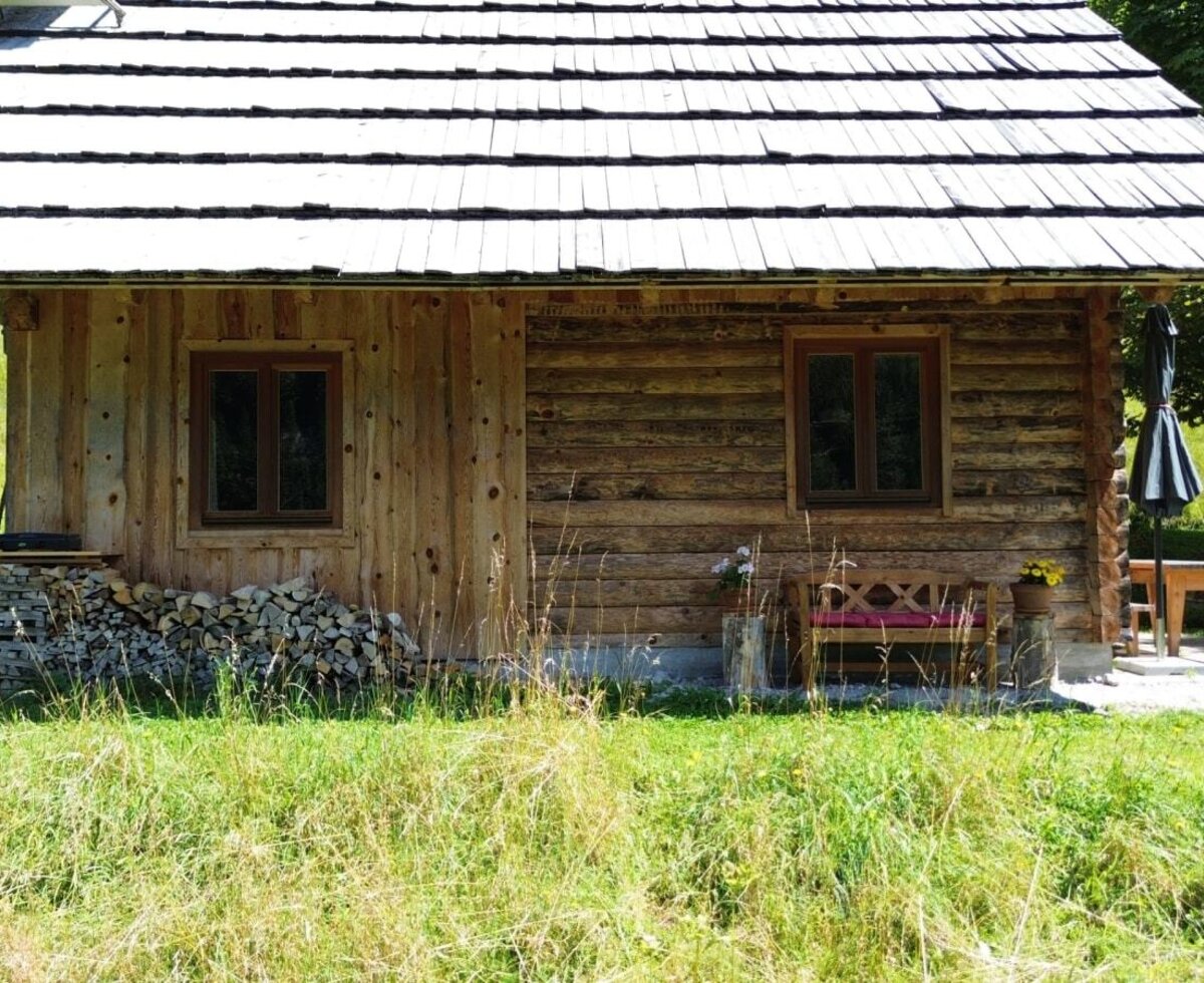 Das rustikale Almhütte mit Schindeldach, zwei Fenstern, gestapeltem Brennholz und einer Terrasse mit Tisch und Sonnenschirm.